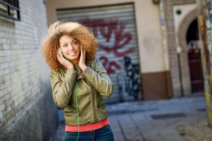 Woman with curly hair and green jacket pictured using headphones in backstreet