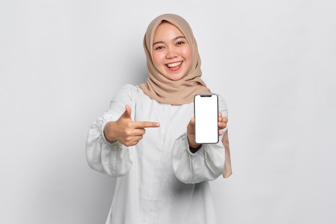 Asian Muslim woman in a bright studio shoot holding cell phone and pointing towards phone