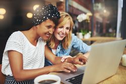 Two female friends sitting at a cafe table smiling at something on the laptop