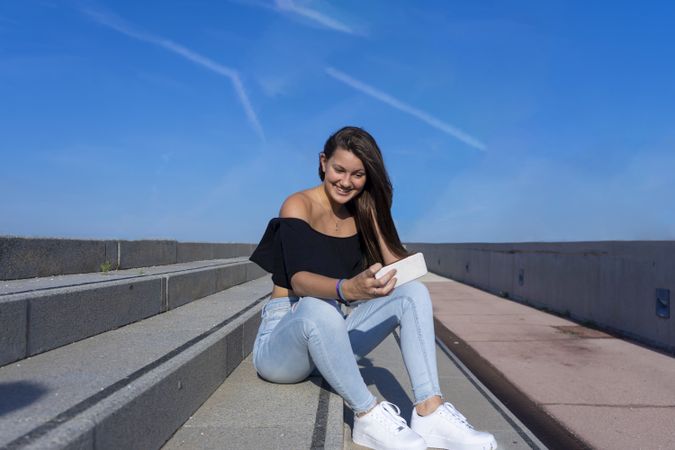 One woman with long hair sitting on staircase while using mobile phone with a big smile