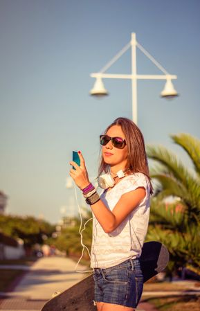 Young girl with skateboard and headphones looking smartphone