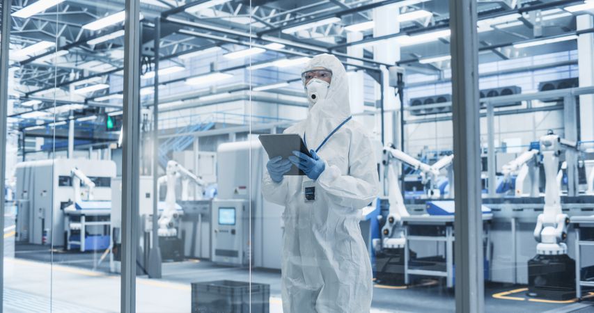 Portrait of Female Worker Wearing Full-Body Protective Coveralls, Mask and Goggles at a Biotechnology Manufacturing Plant. Young Specialist Using a Tablet Computer, Overlooking Nanotech Production