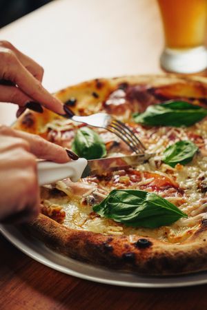 Person slicing hot Italian pizza with fresh basil and melted cheese
