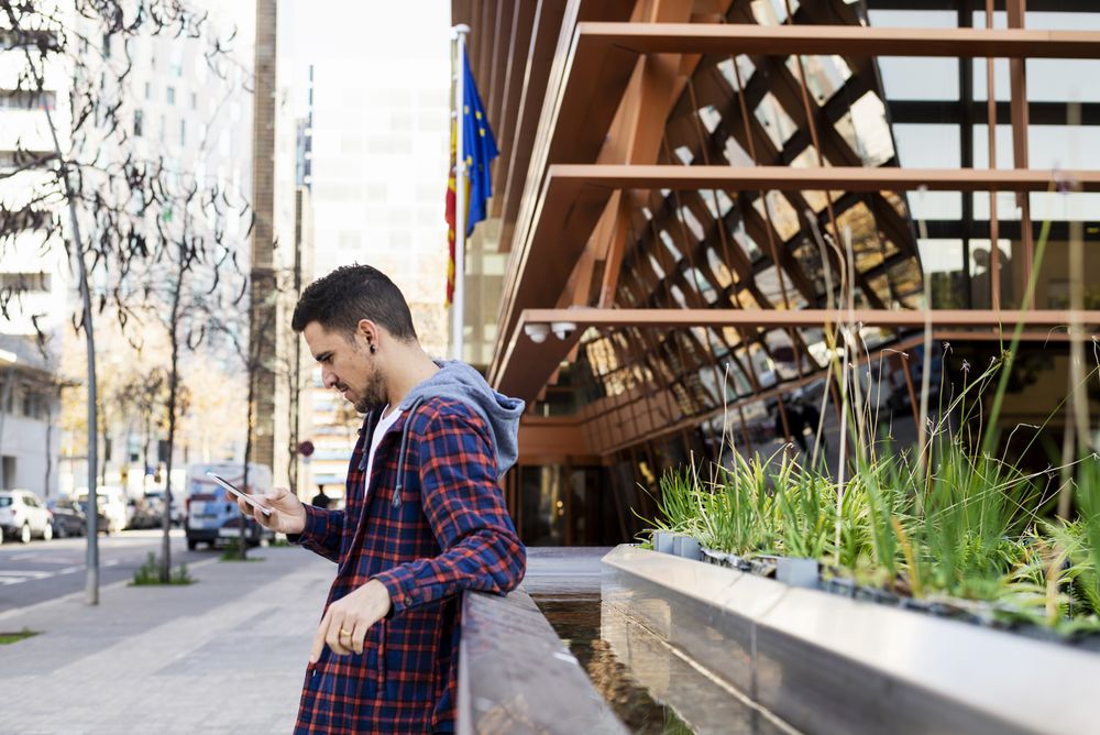 Side view of man wearing scarf leaning on metallic fence while using smartphone outdoors