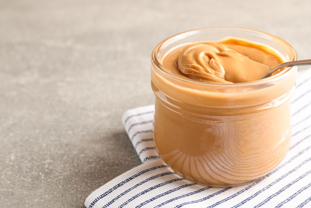 Glass jar with peanut butter and spoon, and kitchen towel on grey background, space for text and closeup
