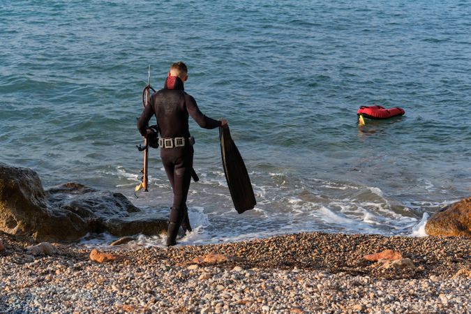 Anonymous man in wetsuit walking in seawater with flippers and speargun