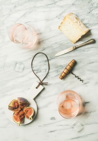 Two glasses of rose wine on marble table with quartered fig, cheese, knife and corkscrew