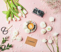 Coffee, camera, tulips artfully arranged on pink background, with “happy” card, square crop