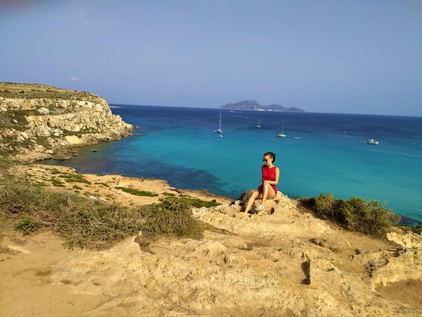 Woman in red dress sitting on a rock near seashore in Sicily, Italy
