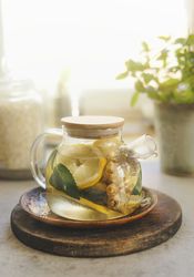 Glass teapot with lemon, mint and ginger on wooden cutting board at window background with natural light