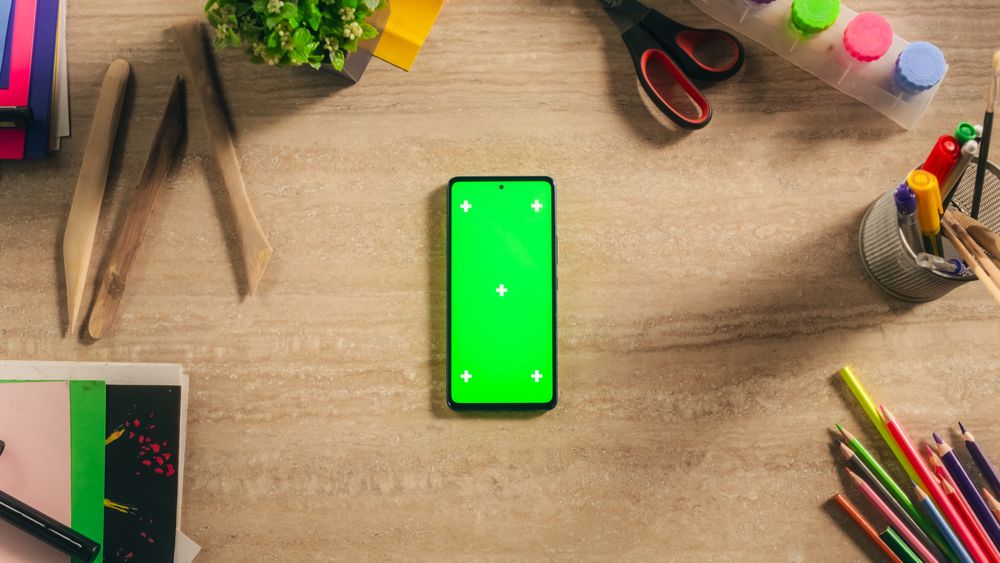 Top Down View of a Smartphone with Mock Up Green Screen Chromakey Display with Motion Tracker Placeholders. Cellphone Device on a Table with Designer Art Supplies and Tools. Social Media Promotion