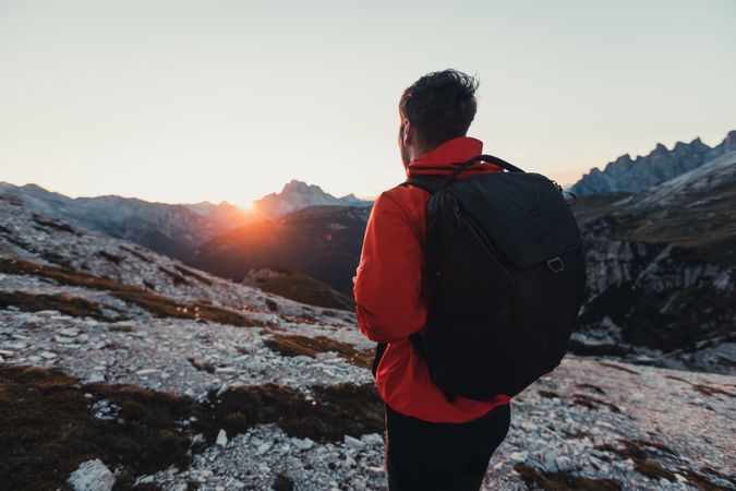 Back view of man in red jacket with backpack gazing the horizon in the mountains at sunset