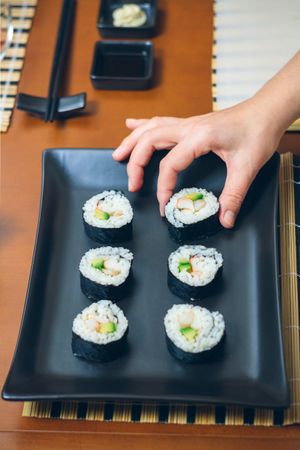 Hand of female chef placing Japanese sushi rolls with rice, avocado and prawns on nori seaweed sheet arranged on tray