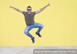 Smiling male jumping in front of yellow wall with outstretched arms bGLra0