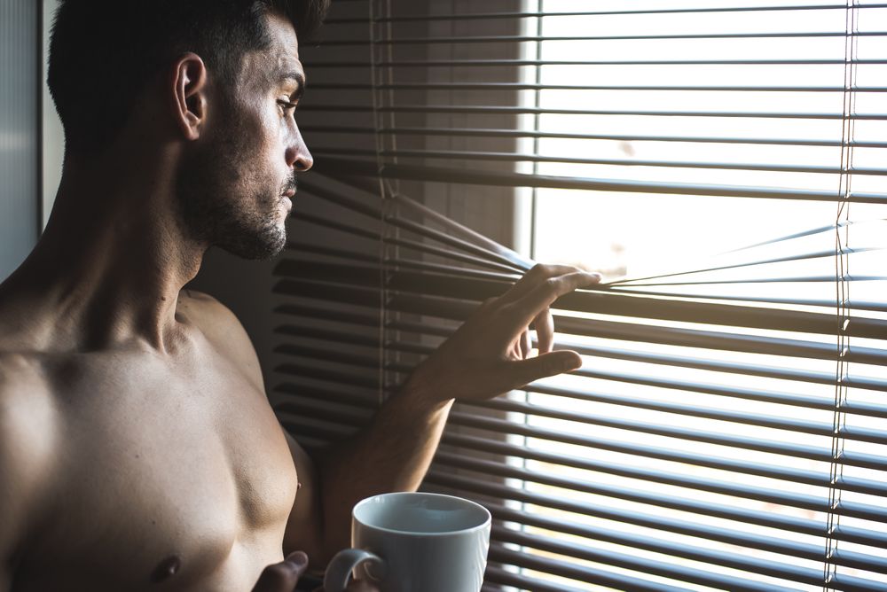 Man standing by a windows blind with a cup of coffee