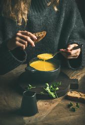 Blonde woman eating warm yellow soup from dark bowl holding toast