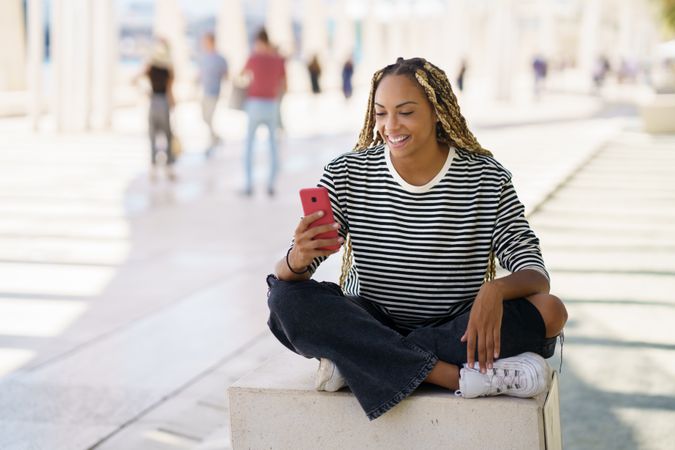 Woman smiling and checking mobile phone while sitting in pedestrian area outside