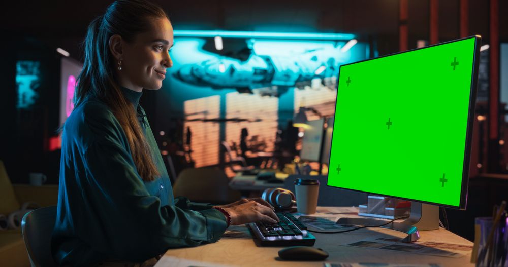 Portrait of Young Woman Sitting at Her Desk Using Desktop Computer with Mock-up Green Screen.
