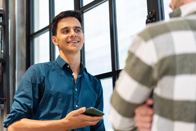 Smiling male employee in shirt holding smartphone chatting with coworker
