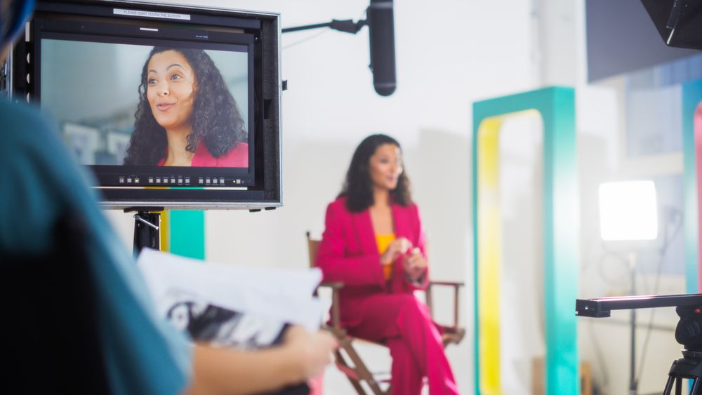 Vibrant On-set Image Of A Young Female Host In A Pink Suit, Engaging In A Lively Discussion During A Tv Show Recording, Surrounded By Modern Videography Equipment And Colorful Set Design.