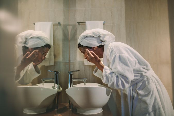 Side view of woman in bathrobe washing her face in bathroom sink and looking into the mirror