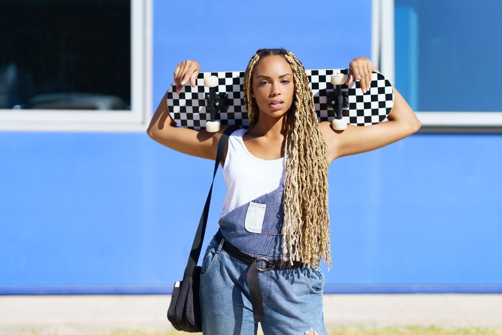 Female in denim overalls standing with skateboard behind head in front of blue building