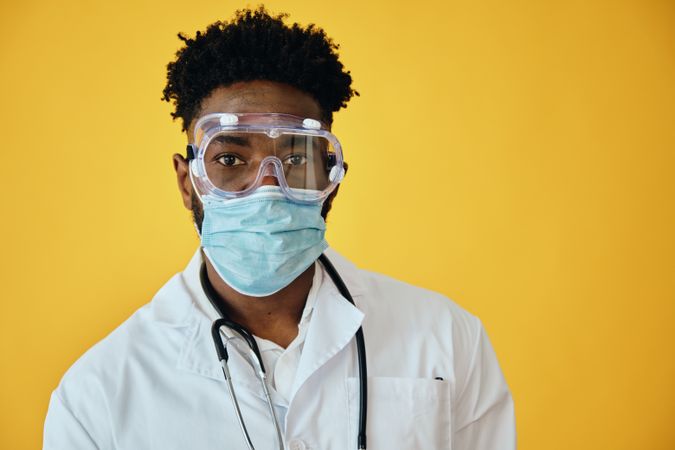 Portrait of Black male doctor in yellow studio with stethoscope, face mask and eye goggles