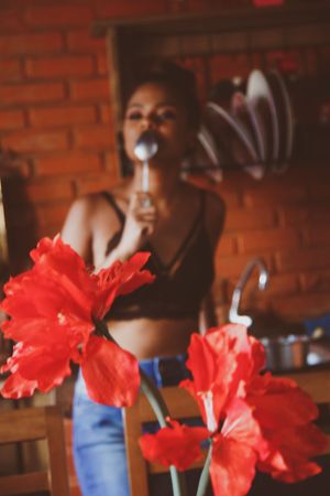 Woman in dark bra eating with spoon standing near red flowers in the kitchen