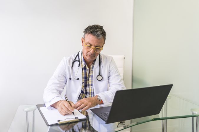 Male doctor working on laptop and clipboard in medical office