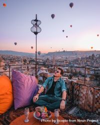 Man sitting on cushion on roof of building under sky filled with hot air balloon 0PWPlb