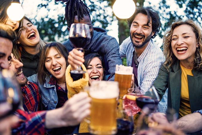 Joyous multi-ethnic friends toast with beer, wine, and sangria at an outdoor party at dusk under garden lights