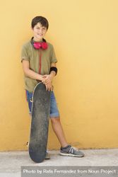 Young boy leaning on a yellow wall with headphones on neck, holding a skateboard 49ml8B