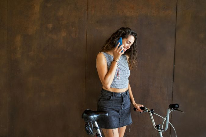 Smiling woman talking on cell phone leaning on bike in front of wall