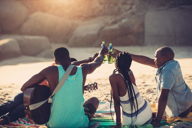 Rear view of young people toasting with beer bottles while sitting on beach