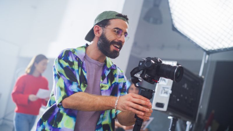 Young Male Cinematographer With Beard, Wearing A Colorful Jacket And Cap, Operates A Camera In A Studio Setting, With A Female Assistant In Background, Focusing On A Creative Shoot.