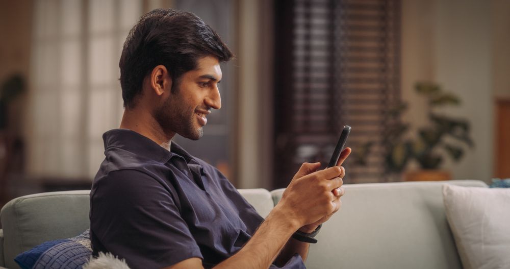 Indian Man Using Smartphone at Home: Seamlessly Managing Personal Finances, Exploring New Recipes