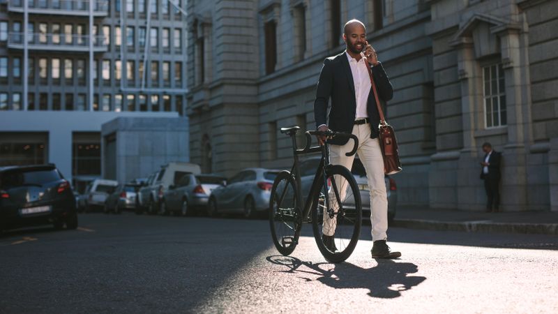 Young businessman walking outdoors with a bicycle and using mobile phone