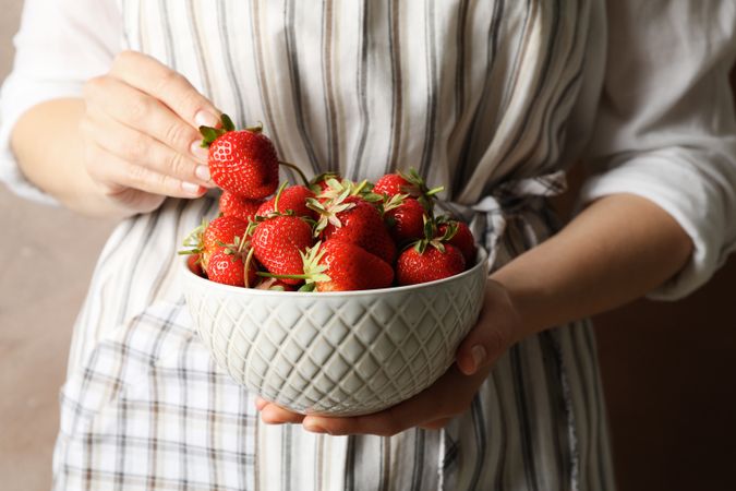 Woman holds bowl with tasty strawberry against brown background, close up.