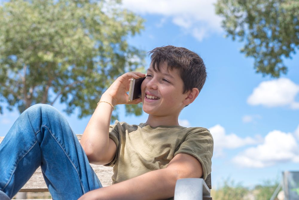 Smiling boy sitting on bench speaking on smartphone