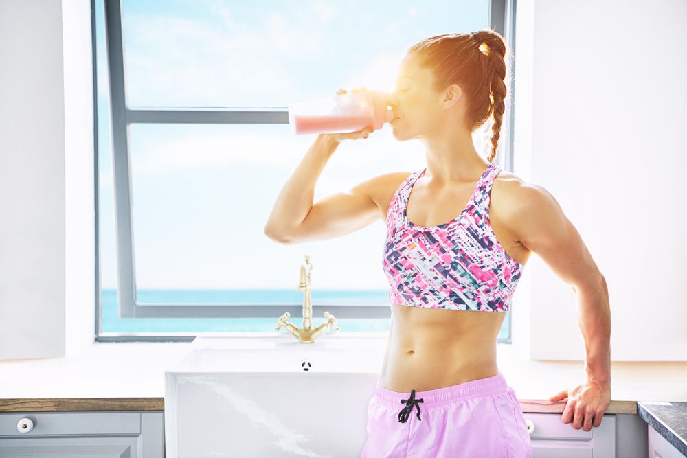 Muscular woman standing by bright kitchen window drinking fresh fruit smoothie