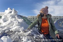 Woman with facemask removing snow off her car's windshield 0LAZR5