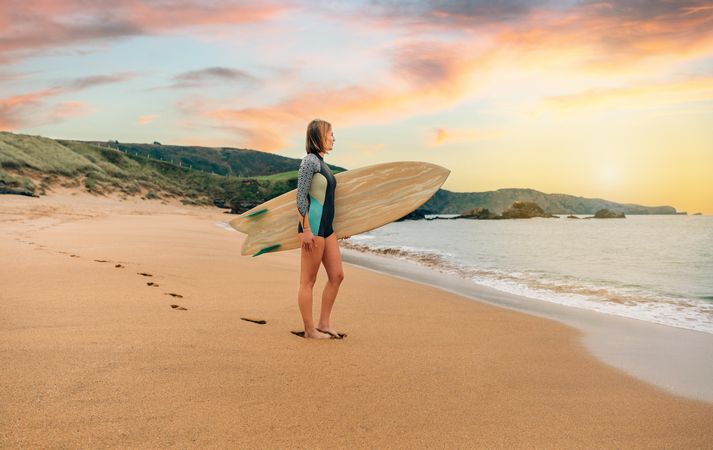 Woman standing on sand with surfboard gazing out at beautiful ocean view