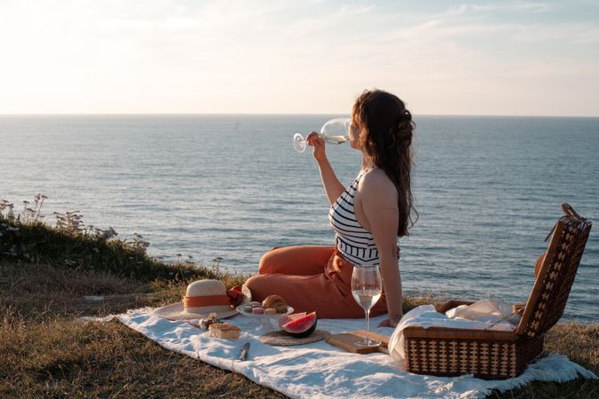 Woman with glass of drink on picnic mat looking at sea and mountains