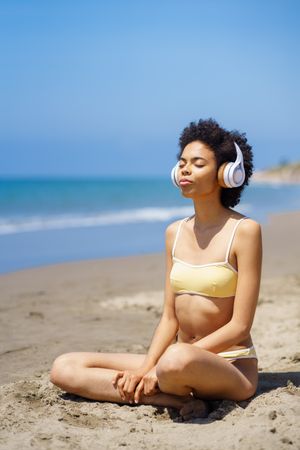 Calm woman in yellow swimwear sitting and meditating on beach looking out to the ocean, vertical