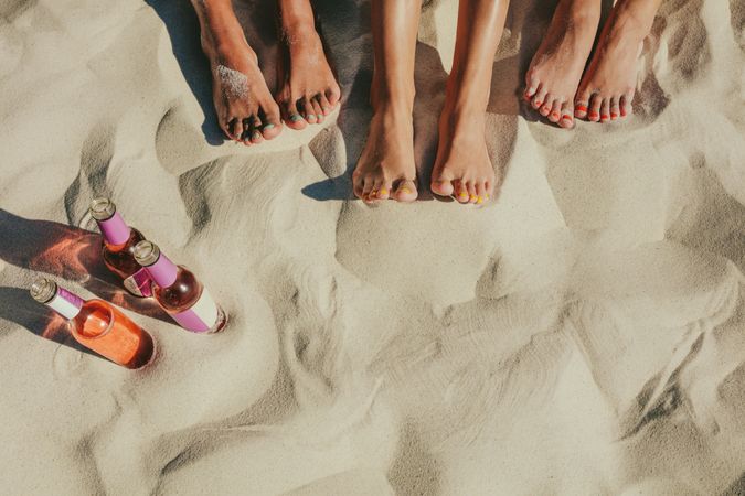 Feet of group of females in sand with glass bottles to the side