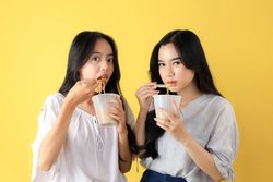 Two Young Asian Girl Holding A Cup of Instant Noodle, Isolated on Yellow Background.
