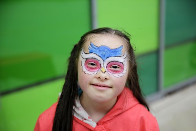 Adorable girl with Down syndrome with face paint standing in front of green wall