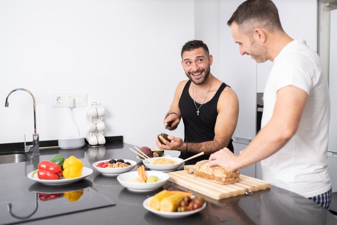 Smiling male couple preparing vegetables and cutting bread for lunch in bright kitchen