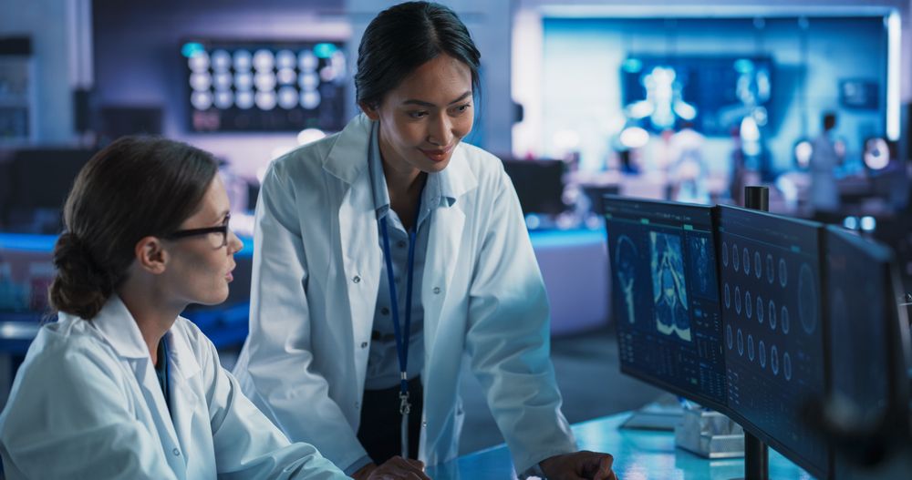 Medical Research Center Laboratory: White And Asian Female Scientists Talking And Using Desktop Computer With MRI Scans Of Human Brain.