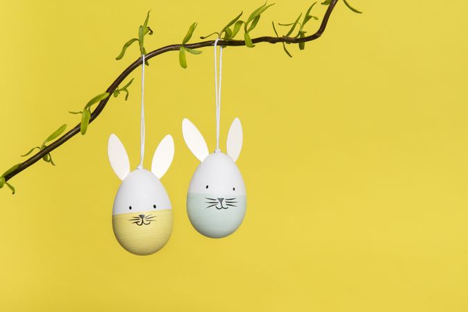 Two easter egg ornaments with happy bunny faces hanging on a branch on a yellow background